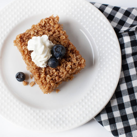 Square of crumble dessert with whipped cream and blueberries on a white plate, checkered cloth in the background